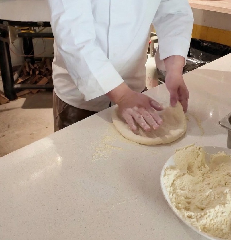 Manos de un chef estirando una bola de masa de pizza cruda sobre una superficie enharinada frente a un horno de leña.