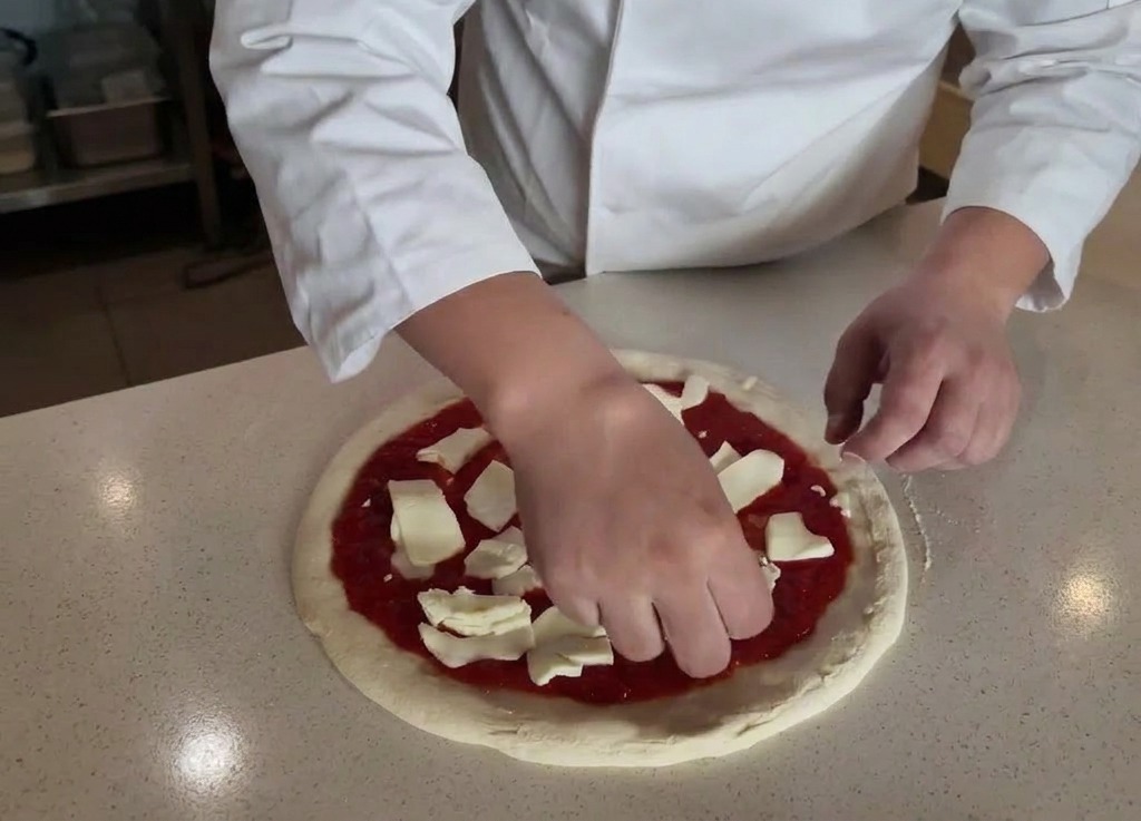 Manos colocando trozos de queso mozzarella blanco sobre la base de pizza cubierta con salsa roja.