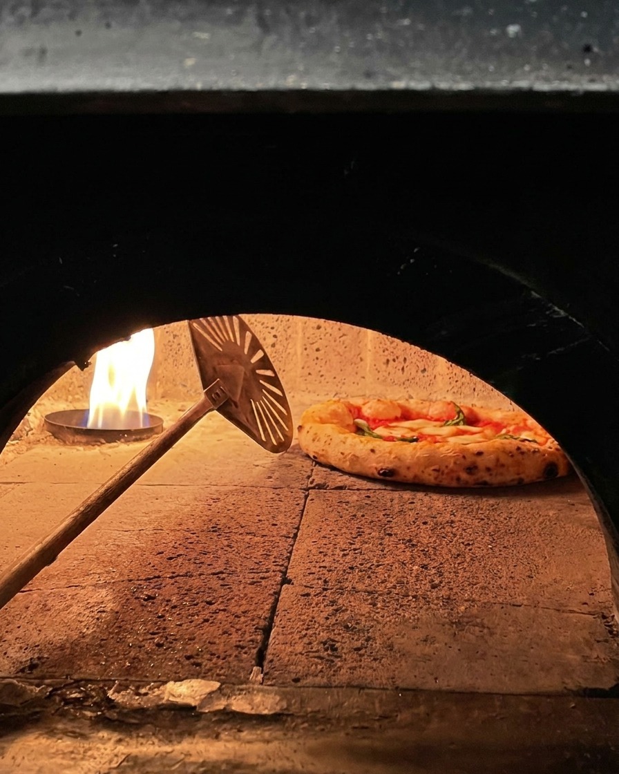 Una pizza Margarita cocinándose dentro de un horno de piedra y siendo girada con una pequeña pala metálica.