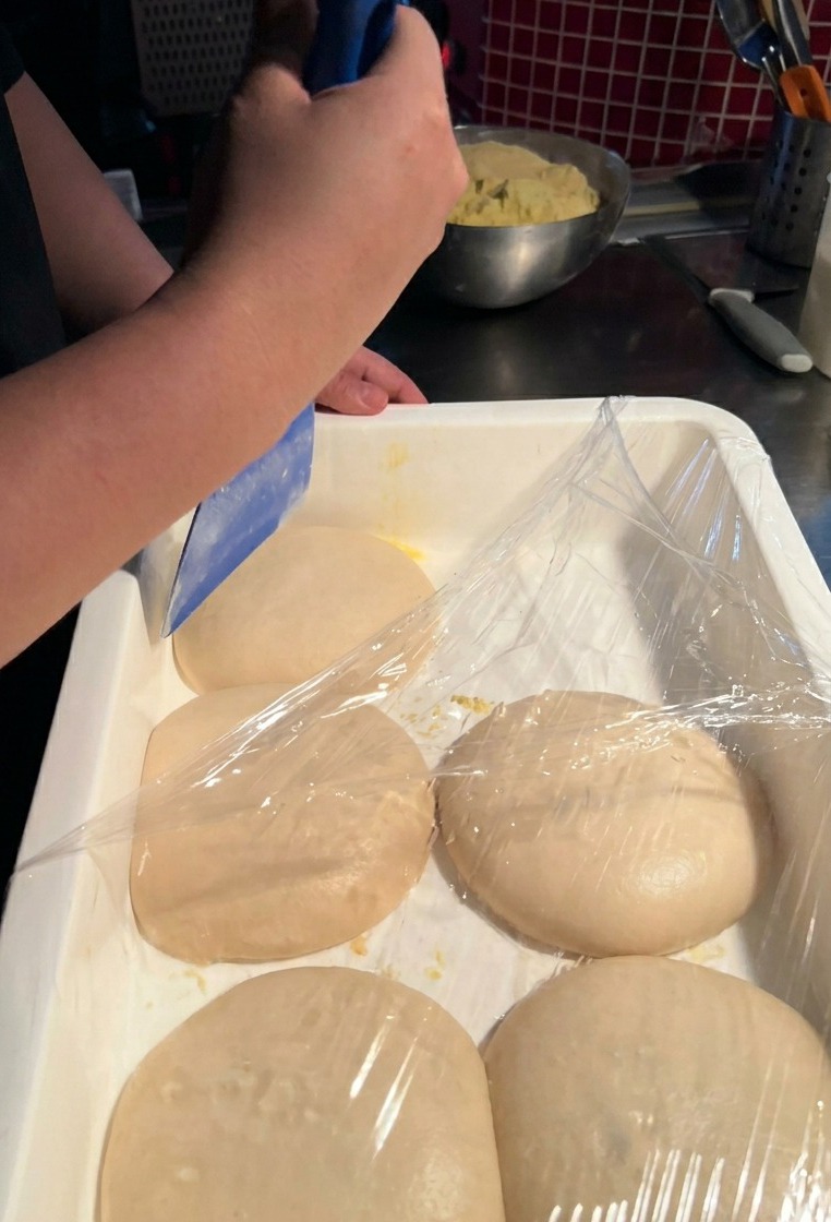 Una persona usando una espátula de plástico azul para levantar con cuidado una porción de masa de pizza de un contenedor blanco.