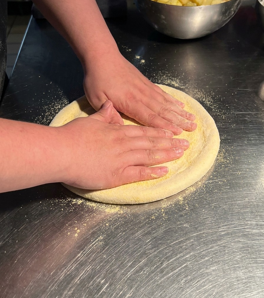 Manos presionando sobre una pieza circular de masa de pizza en una mesa de acero inoxidable, estirándola en un disco más grande.