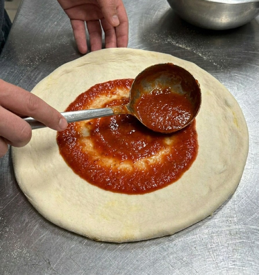 Un cucharón metálico extendiendo salsa de tomate roja en movimiento circular sobre una masa de pizza plana.
