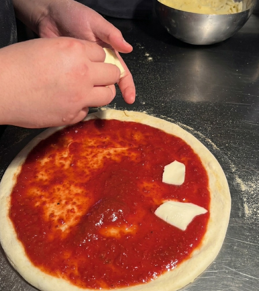 Una persona colocando trozos de queso mozzarella cortados a mano sobre una base de pizza con salsa roja.