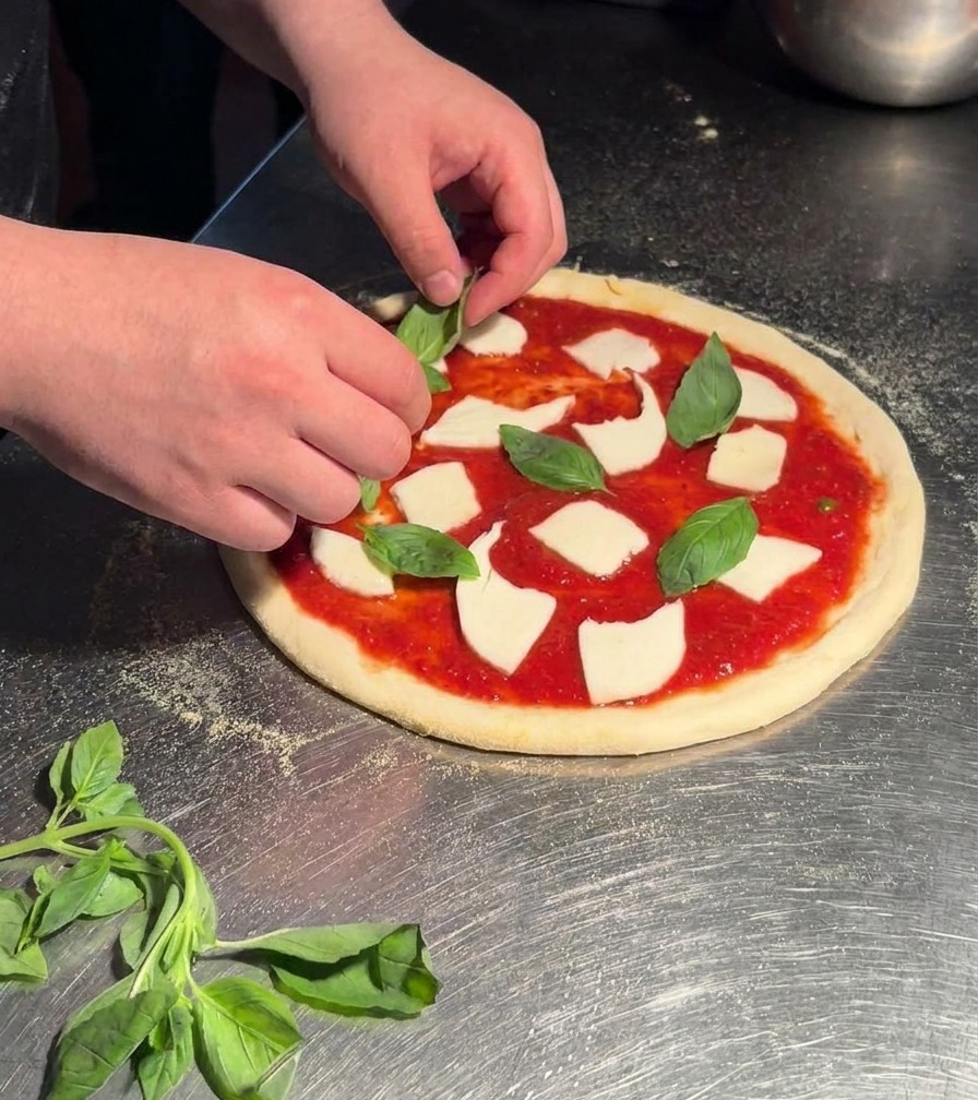 Manos colocando hojas de albahaca verde brillante sobre una pizza Margherita cruda con salsa y mozzarella.