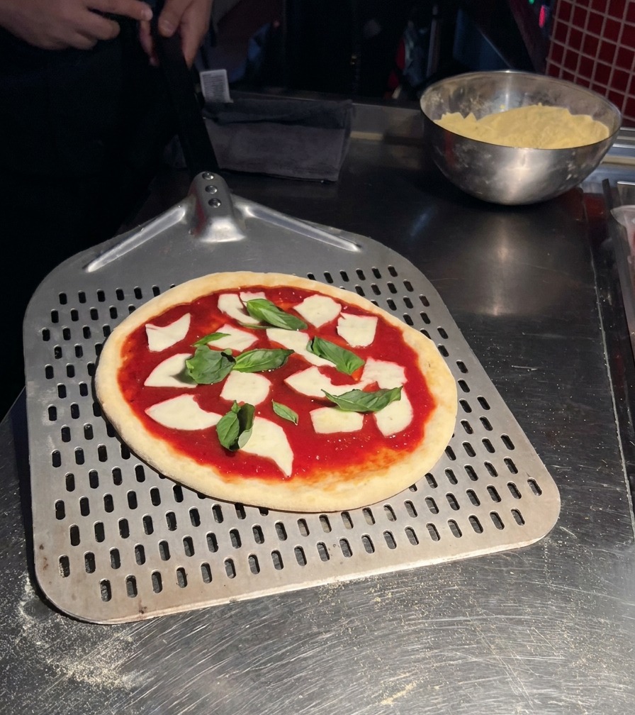 Una pizza Margherita cruda sobre una pala metálica perforada lista para entrar al horno.