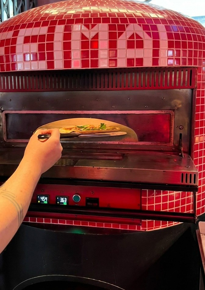 Una persona deslizando una pizza desde una pala metálica hacia el interior de un horno de pizza tradicional de azulejos rojos.