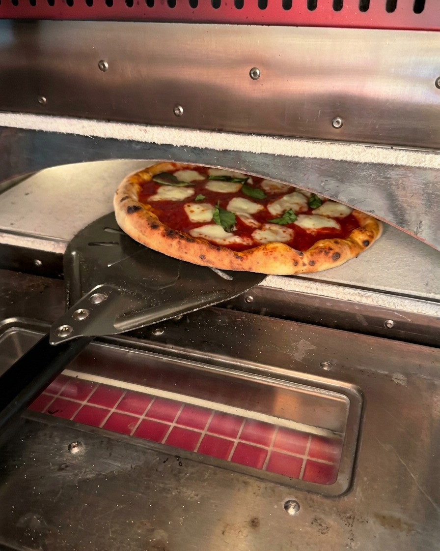 Una pizza recién horneada con bordes tostados siendo retirada del horno con una pala metálica.
