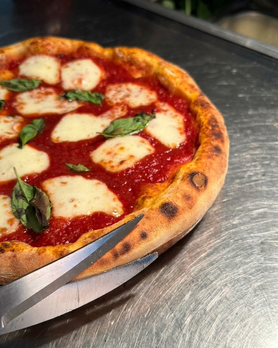Primer plano de unas tijeras metálicas cortando la corteza inflada de una pizza Margherita caliente.
