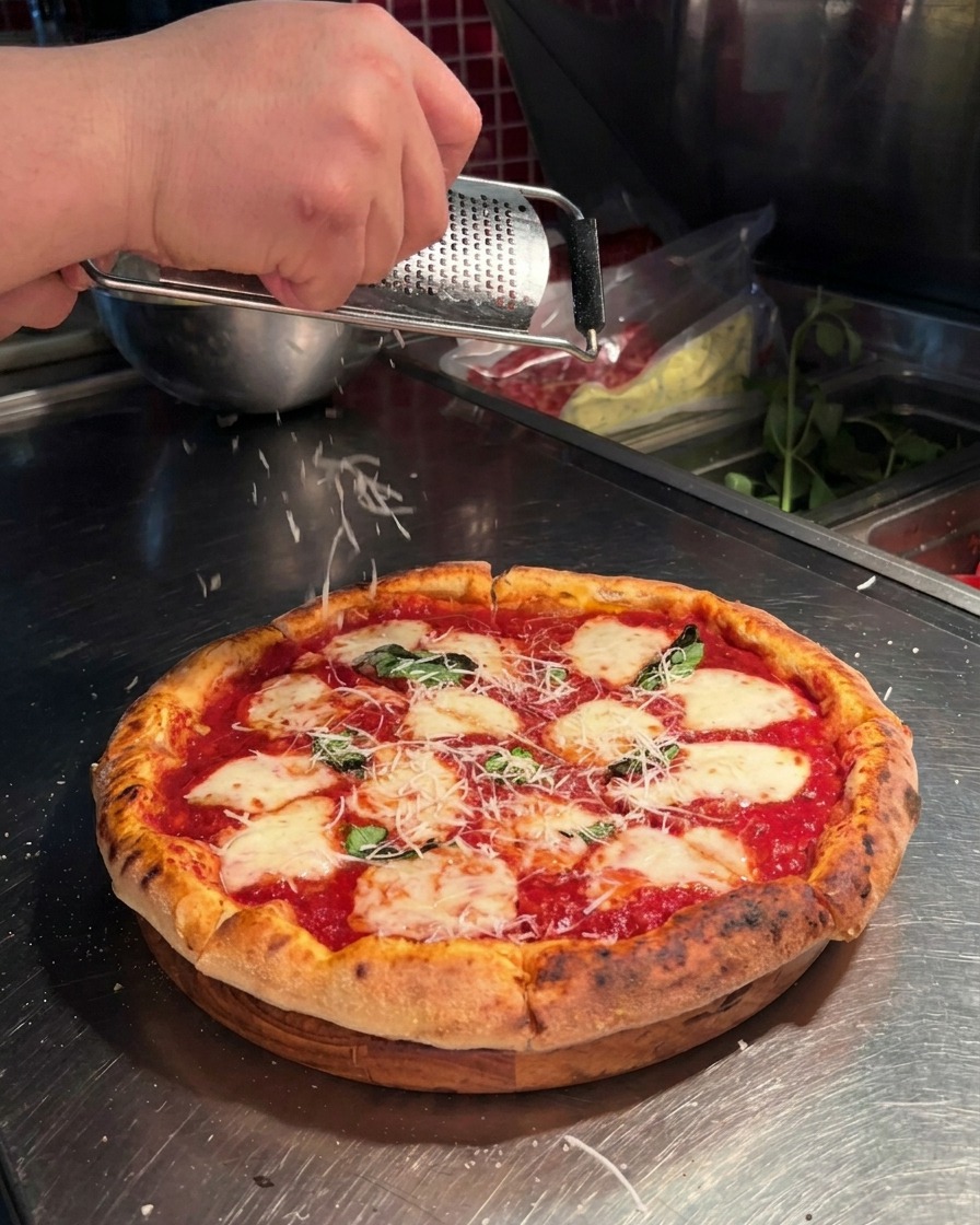 Primer plano de una mano rallando queso parmesano fresco sobre una pizza napolitana.