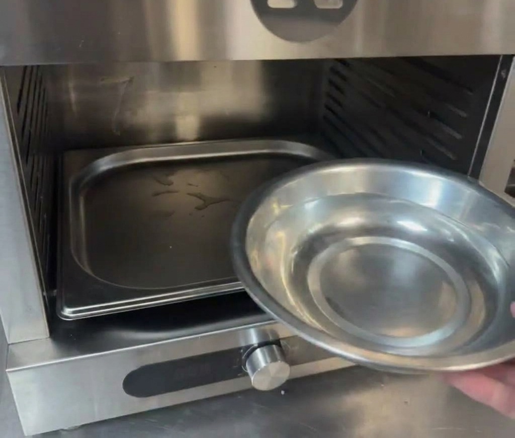 Una mano vertiendo agua desde un cuenco metálico en una bandeja en la base de un horno profesional.