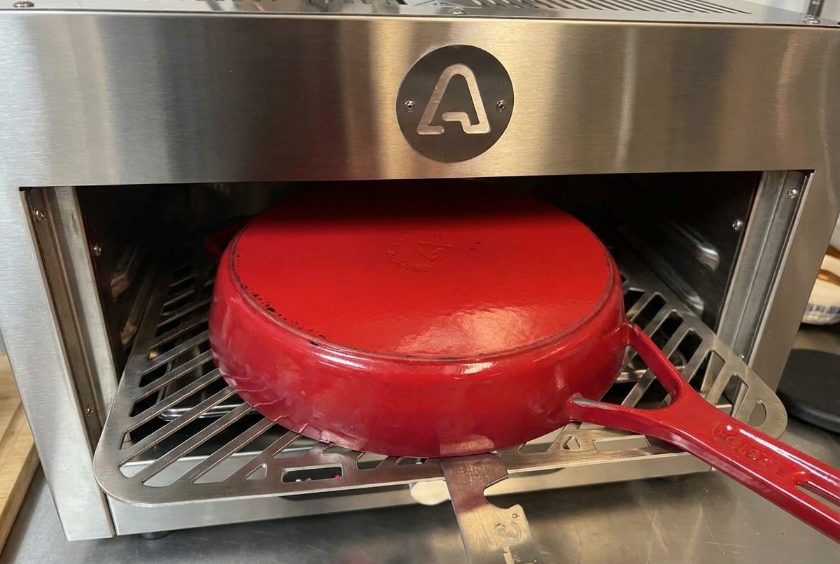 Una olla de hierro fundido roja invertida sobre una rejilla metálica dentro de un horno de acero inoxidable.
