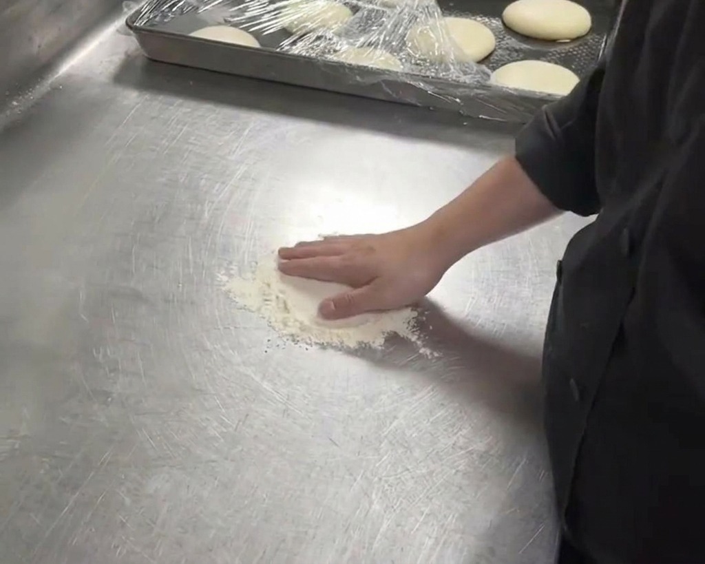 Un chef presionando con la mano una bola de masa blanca sobre una encimera metálica enharinada.