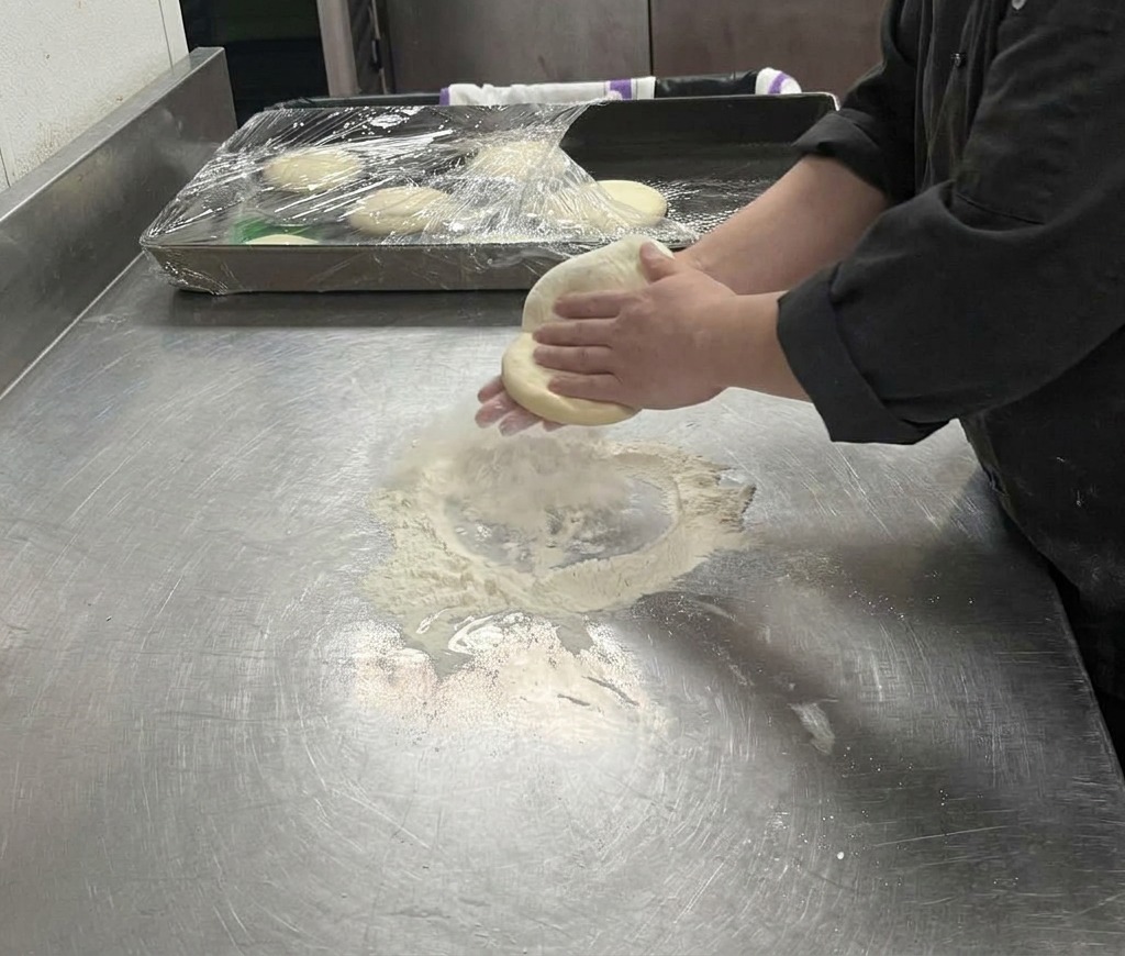 Manos estirando una masa de pizza pálida sobre una superficie enharinada hasta formar un círculo grande.