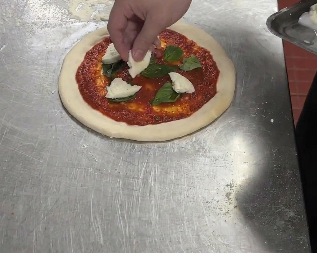 Trozos de mozzarella de búfala blanca siendo añadidos a una pizza con salsa de tomate y albahaca.