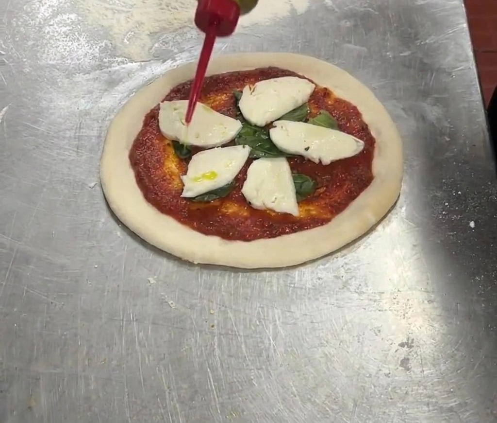 Un biberón rojo vertiendo aceite de oliva virgen extra sobre una pizza con tomate, albahaca y mozzarella.