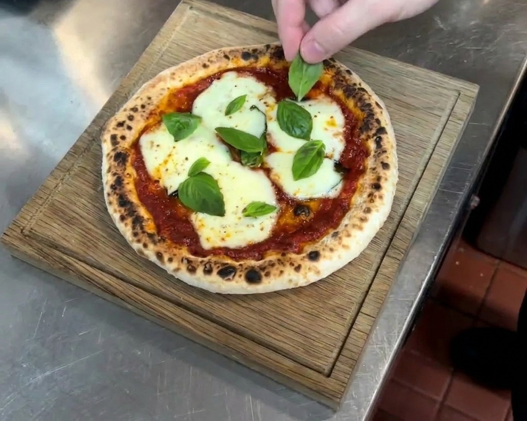 Mano colocando hojas de albahaca fresca sobre una pizza recién horneada con queso fundido y borde moteado.