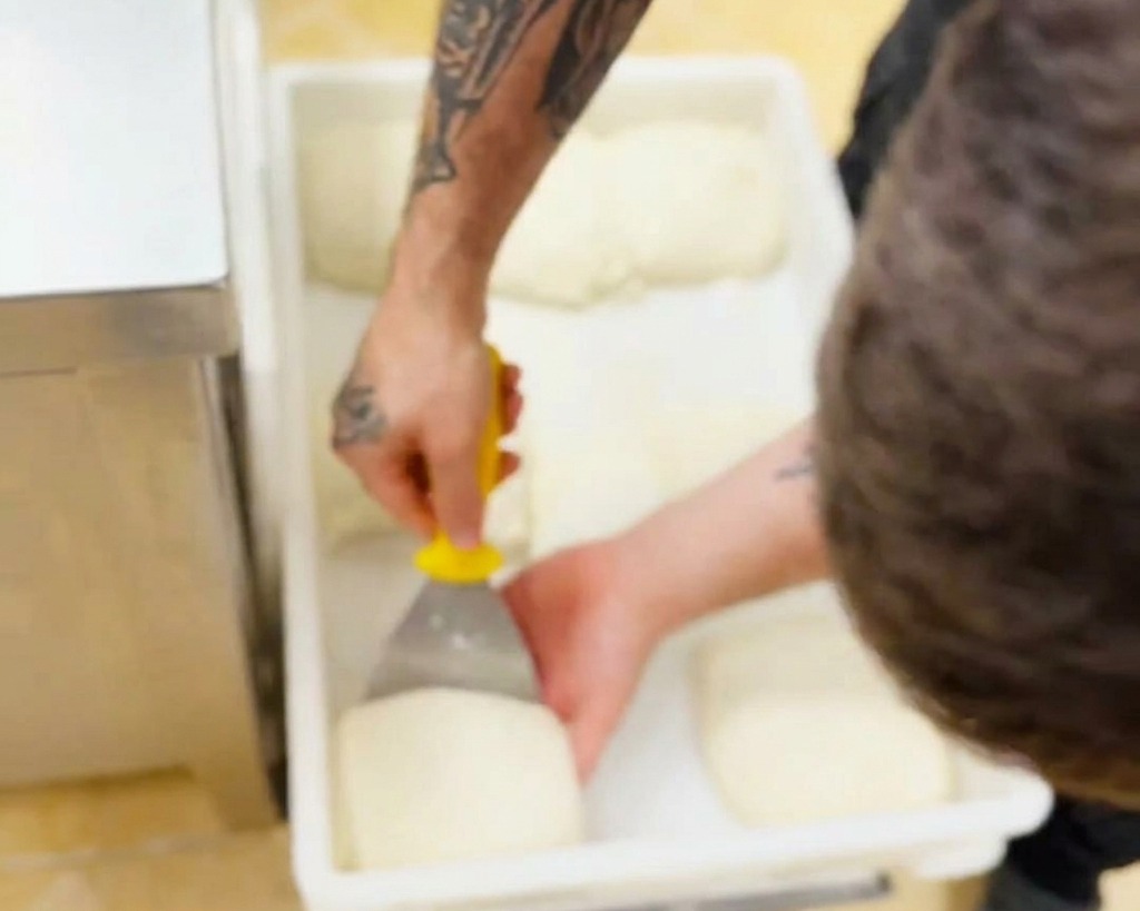Una mano usando un raspador de mango amarillo para levantar con cuidado una bola de masa suave de un contenedor plástico.