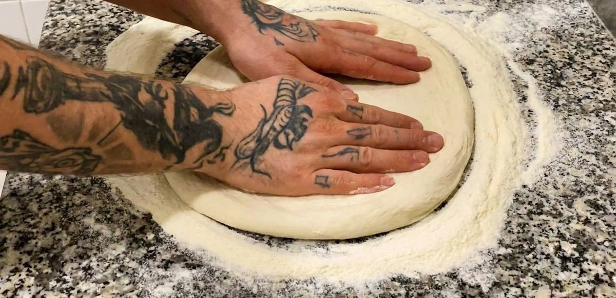 Dos manos presionando una base de pizza redonda y enharinada sobre una encimera de mármol gris.