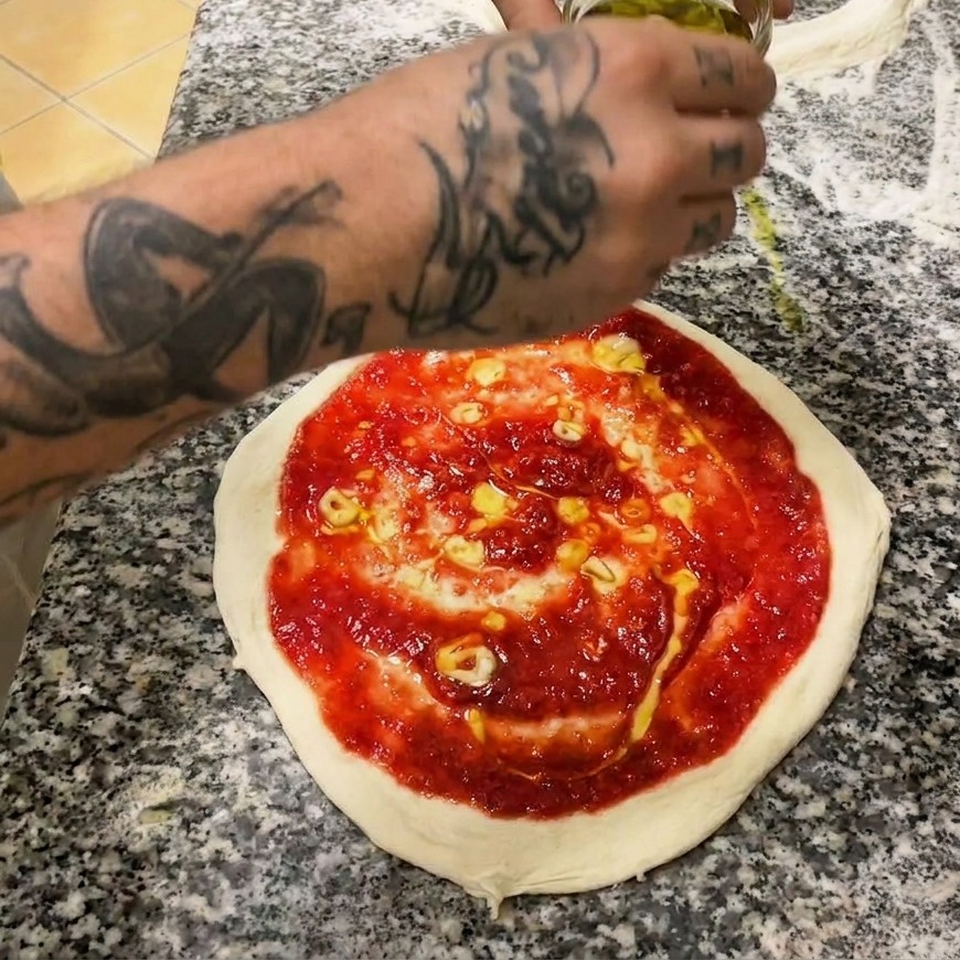 Una mano usando una cuchara metálica para extender salsa de tomate roja en espiral sobre la masa cruda.
