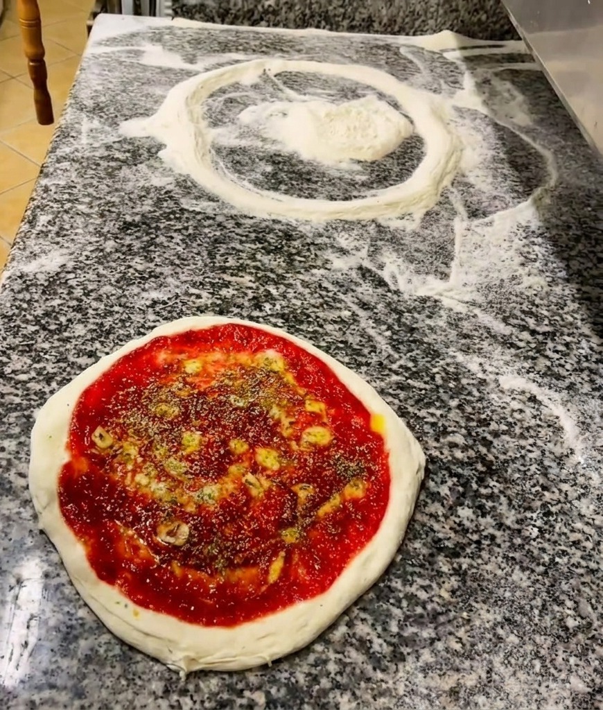 Una mano espolvoreando hierbas secas sobre una base de pizza con salsa de tomate y un chorrito de aceite.