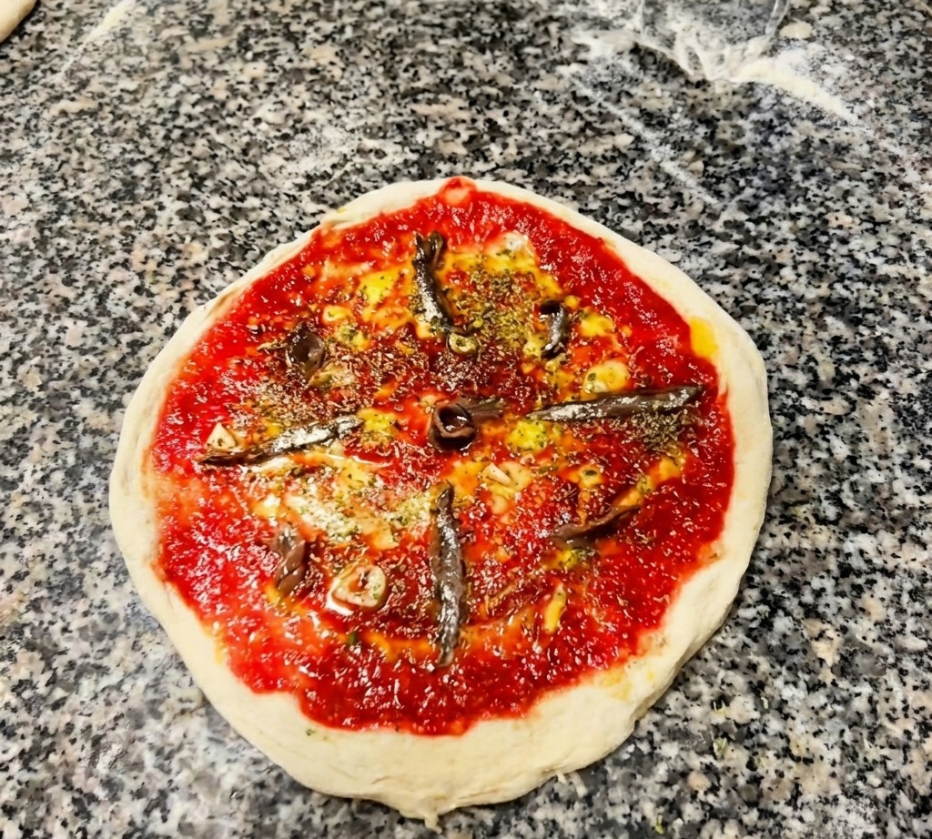 Toma aérea de una pizza cruda con salsa de tomate, hierbas y filetes de anchoa dispuestos de forma radial.