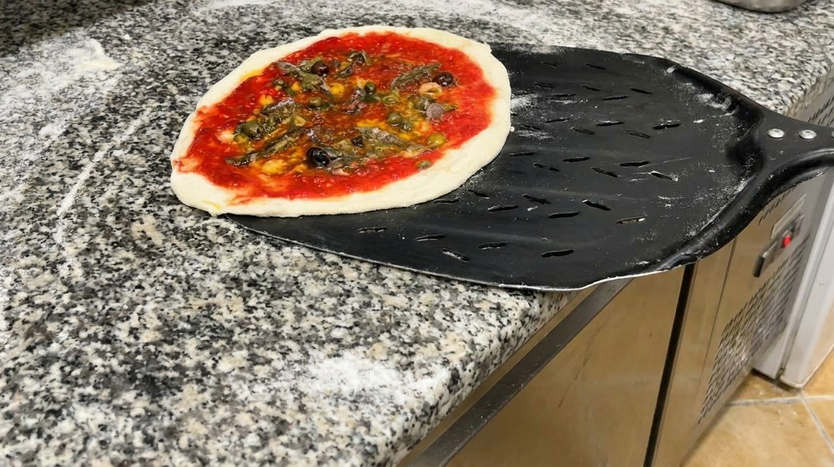 Una pizza de mariscos lista para hornear sobre una pala metálica perforada en una encimera de granito.