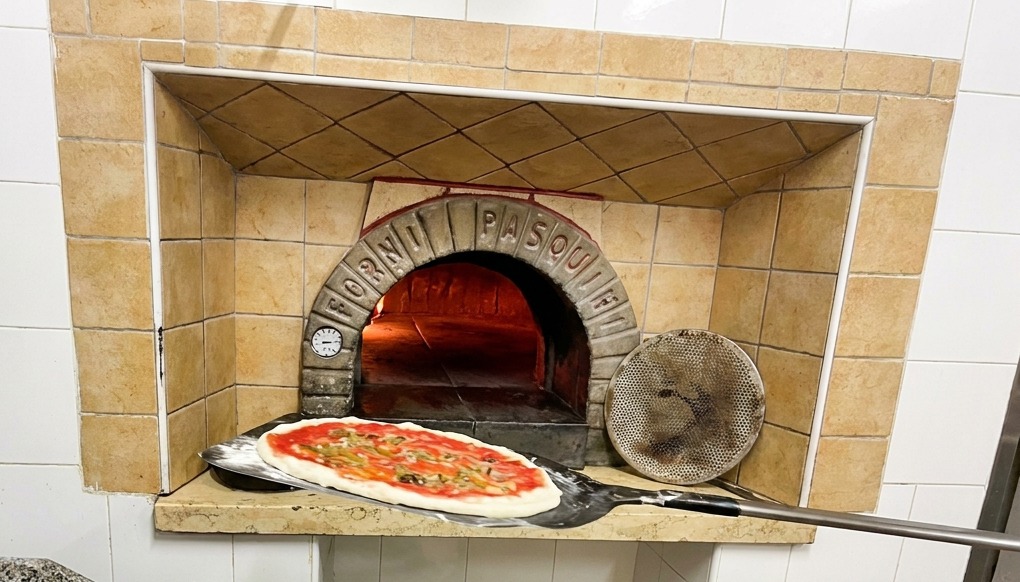 Una pizza en una pala de mango largo siendo introducida en un horno de leña tradicional con llamas vivas.