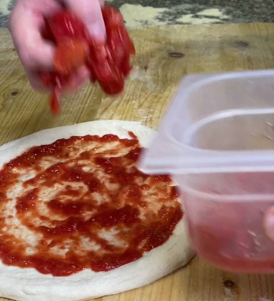 Una mano dejando caer trozos de pulpa de tomate triturado sobre una base de pizza ya cubierta con salsa roja suave.