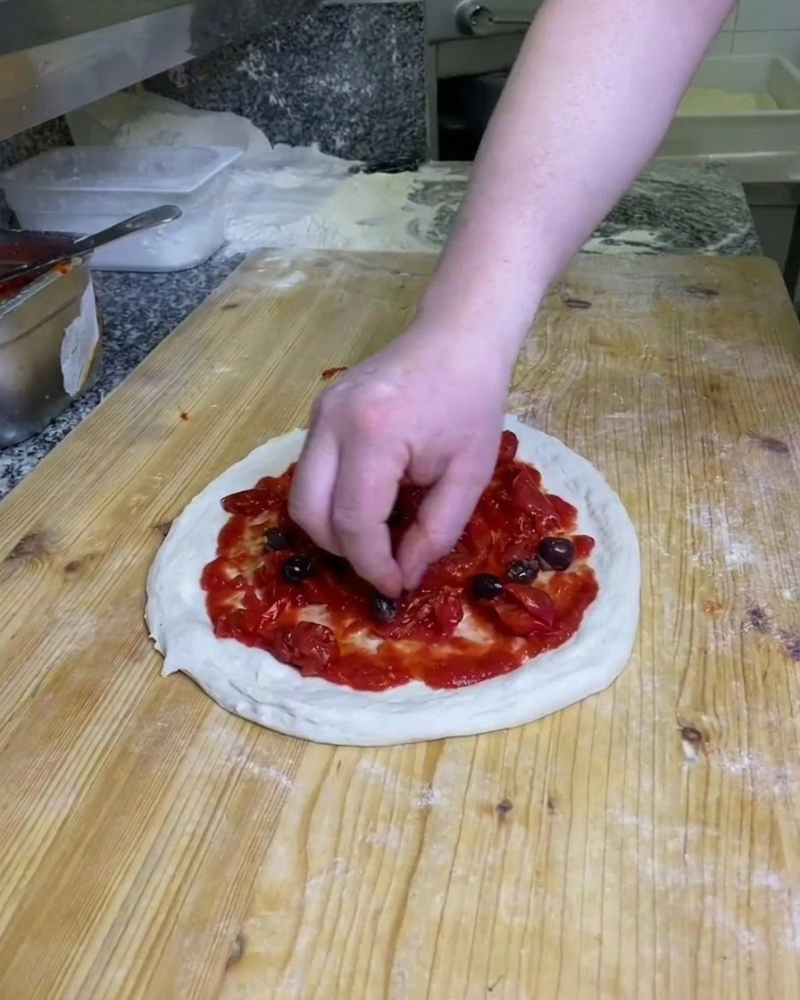 Una mano colocando aceitunas negras enteras sobre la masa de pizza cubierta de tomate en una superficie de madera enharinada.