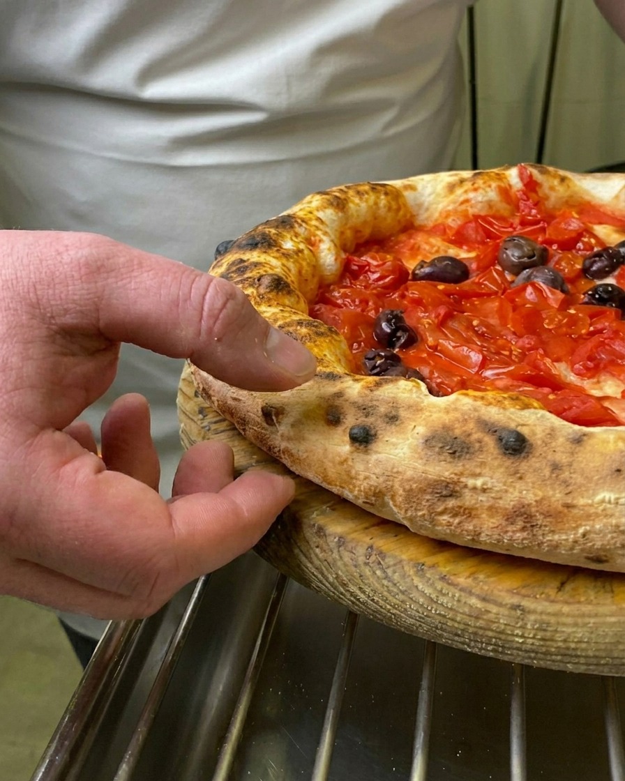 El dedo de un chef presionando el borde inflado y tostado de una pizza Marinara recién horneada.