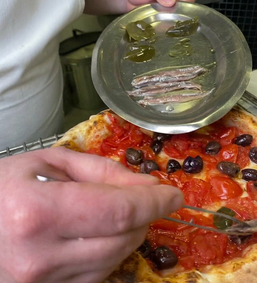 Un chef usando pinzas para colocar una hoja de laurel en escabeche sobre una pizza con tomate y aceitunas.