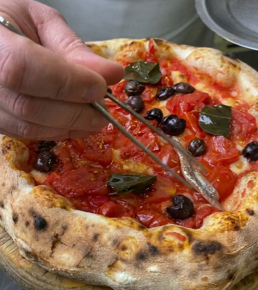 Primer plano de un filete de anchoa plateado siendo colocado sobre la pizza usando pinzas culinarias.