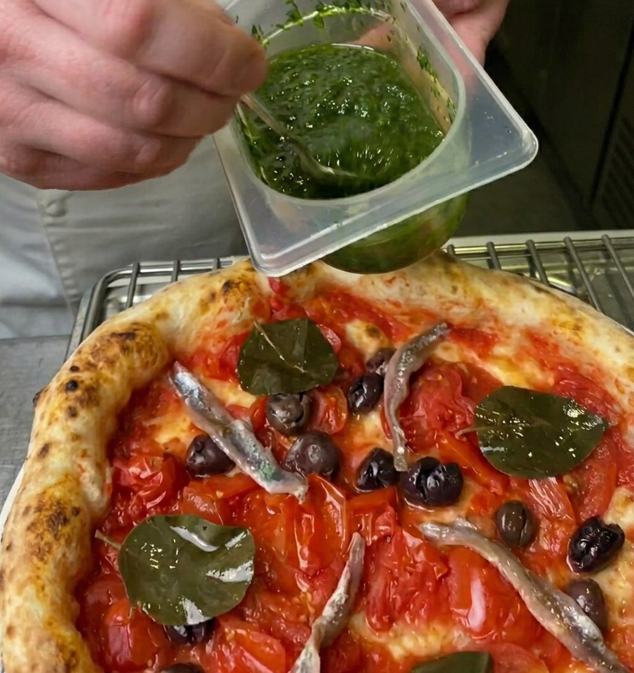 Un chef rociando una salsa de hierbas verde vibrante sobre una pizza usando una cuchara pequeña.