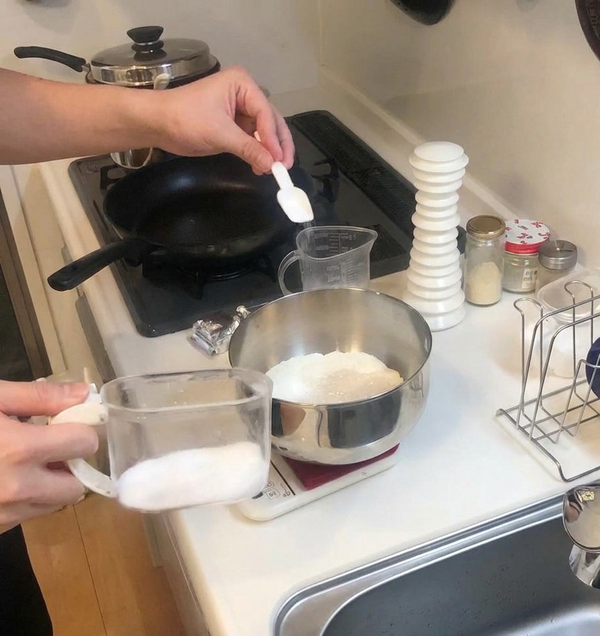 Una pequeña cuchara blanca añadiendo sal a un bol de harina y levadura sobre una báscula de cocina.