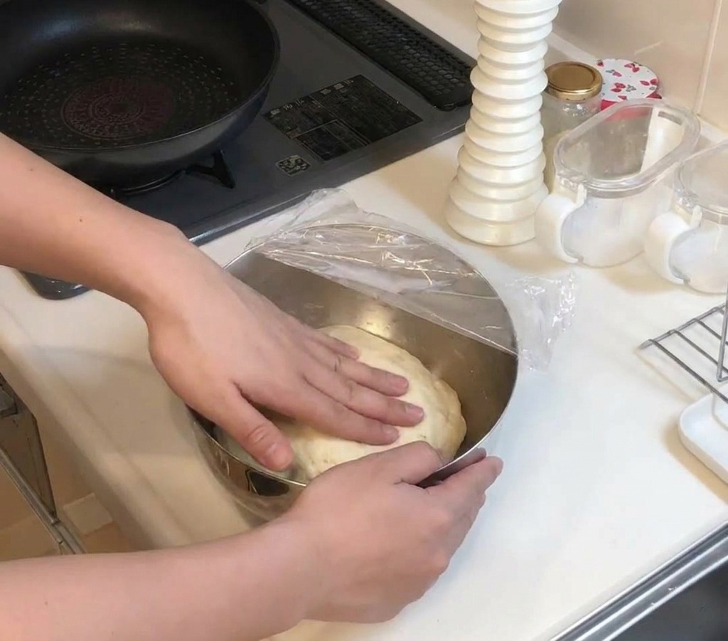 Manos presionando una gran bola de masa pálida que ha subido dentro de un bol cubierto con film transparente.