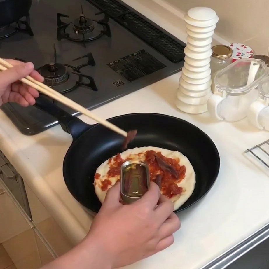 Usando palillos de madera para coger anchoas de una lata metálica y ponerlas sobre la base de pizza con salsa.
