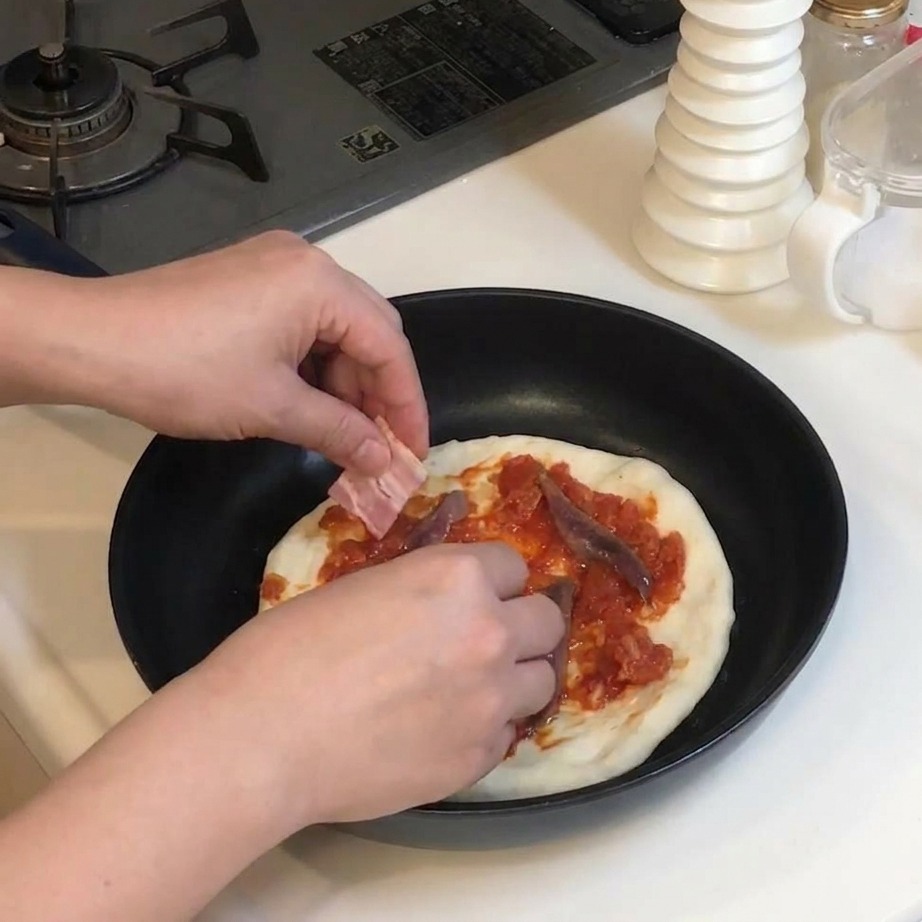 Manos colocando trozos rectangulares de bacón crudo sobre una pizza con tomate y anchoas.