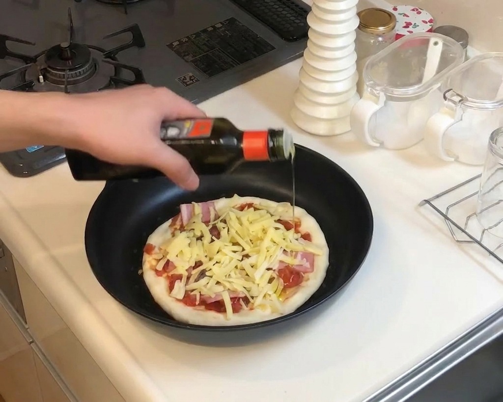 Primer plano de una mano rociando aceite de oliva desde una botella oscura sobre una pizza preparada con queso y bacón en la sartén.