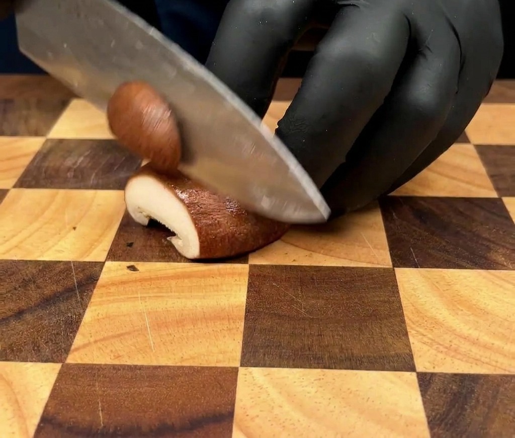 Primer plano de una persona cortando un hongo pleurotus en láminas finas sobre una tabla de madera.