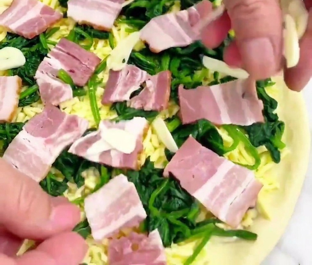 Una mano dejando caer láminas de ajo translúcidas sobre una pizza preparada con espinacas y beicon.