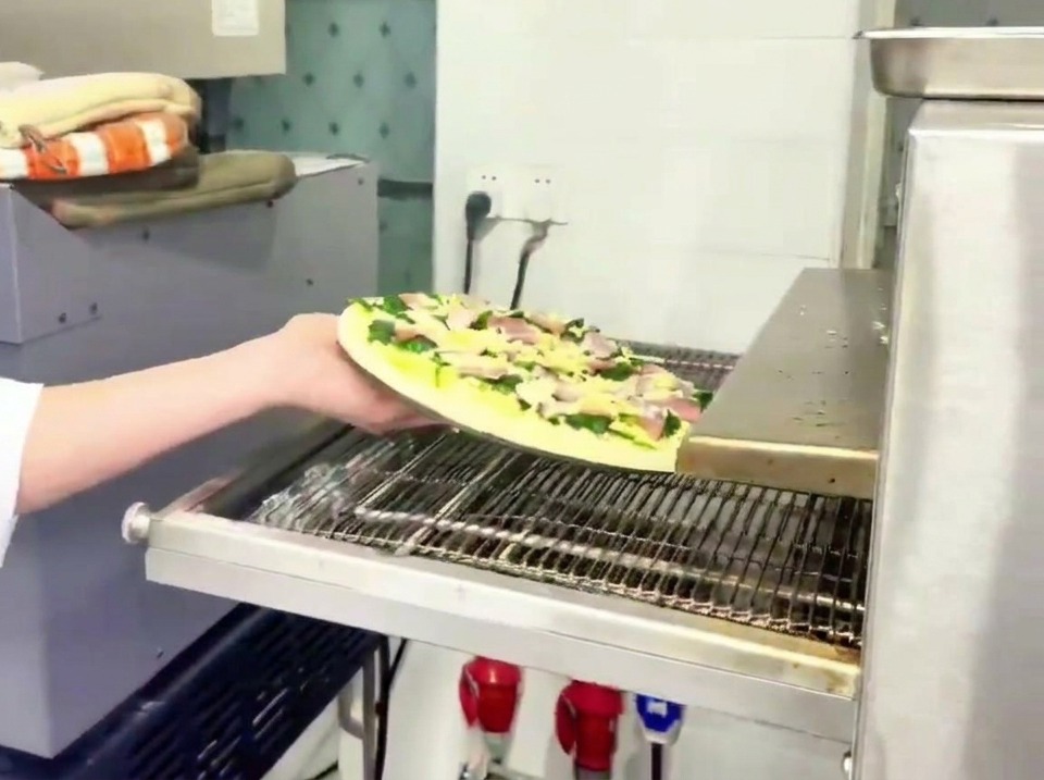 Un operario colocando la pizza de espinacas y beicon en la cinta metálica de un horno profesional.