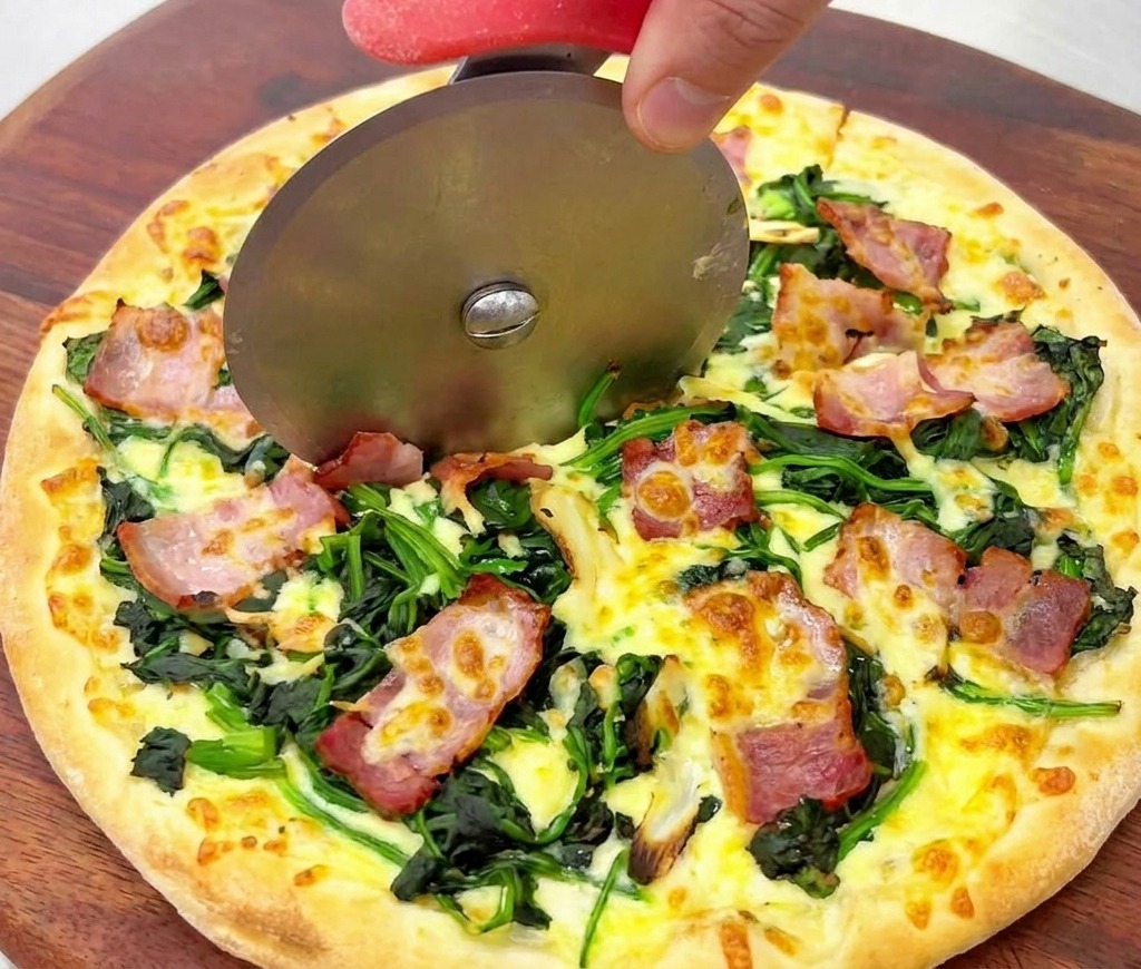 Primer plano de un cortapizzas pasando por una pizza dorada con espinacas y beicon crujiente.