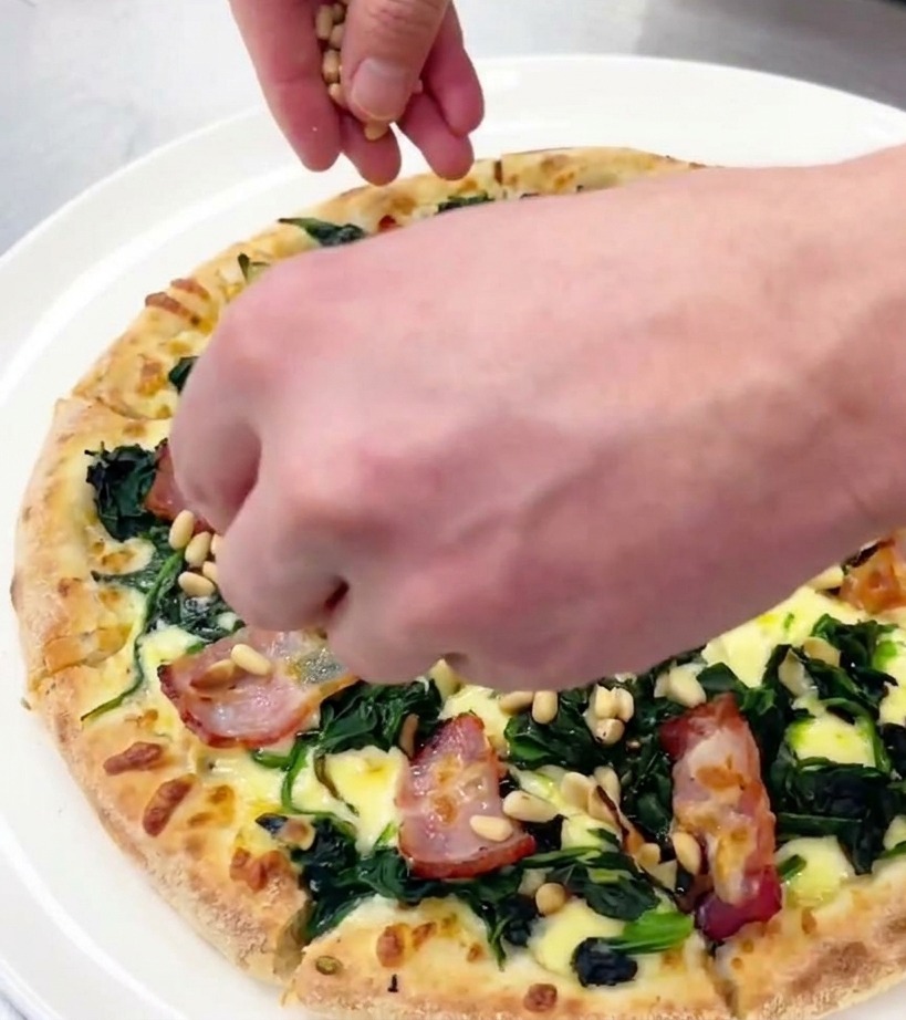 Una mano esparciendo piñones tostados sobre una pizza de espinacas y beicon recién cortada.