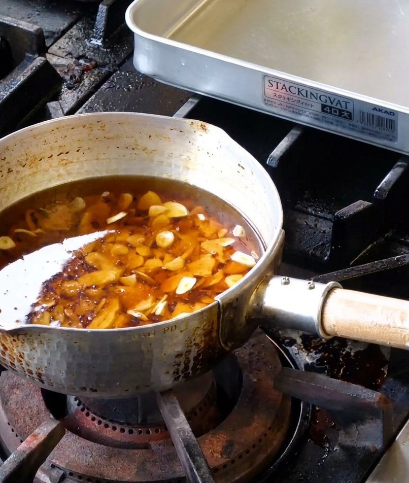 Láminas de ajo crudo sumergidas en una mezcla de manteca líquida y aceite de sésamo en una olla de metal.