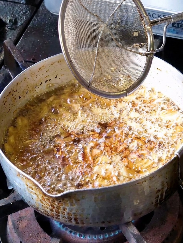 Un colador de malla retirando láminas de ajo frito de color dorado claro de una olla con aceite.