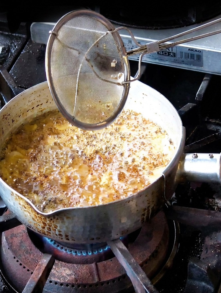 Una mano usando un colador para retirar láminas de ajo frito marrón medio de una olla con aceite oscuro.
