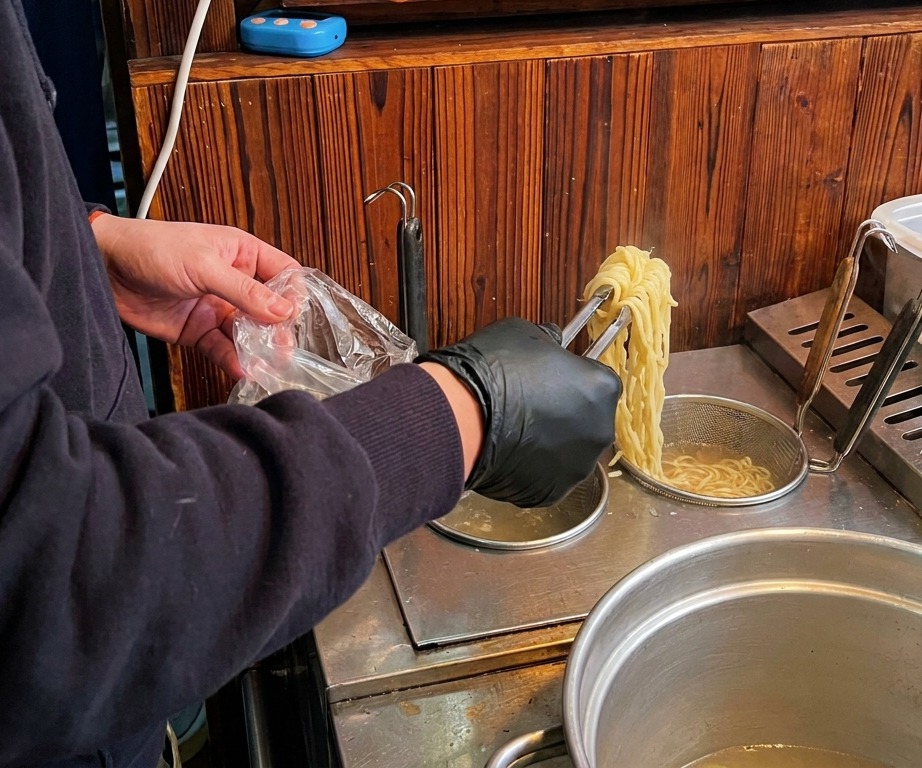 Una persona con guantes negros usando pinzas para levantar fideos ramen largos y amarillos de una olla con agua hirviendo.