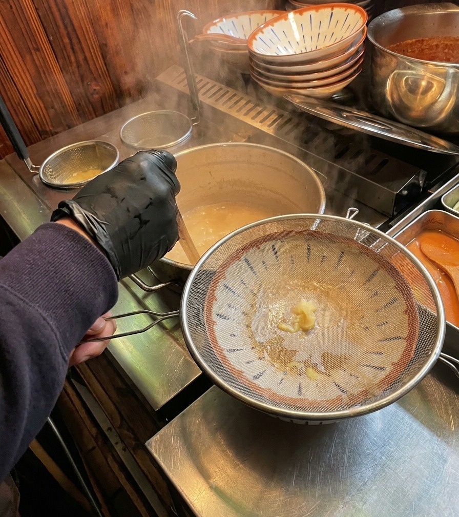 Caldo de huesos blanco y cremoso que sale a través de un colador metálico circular hacia un tazón de ramen decorado.