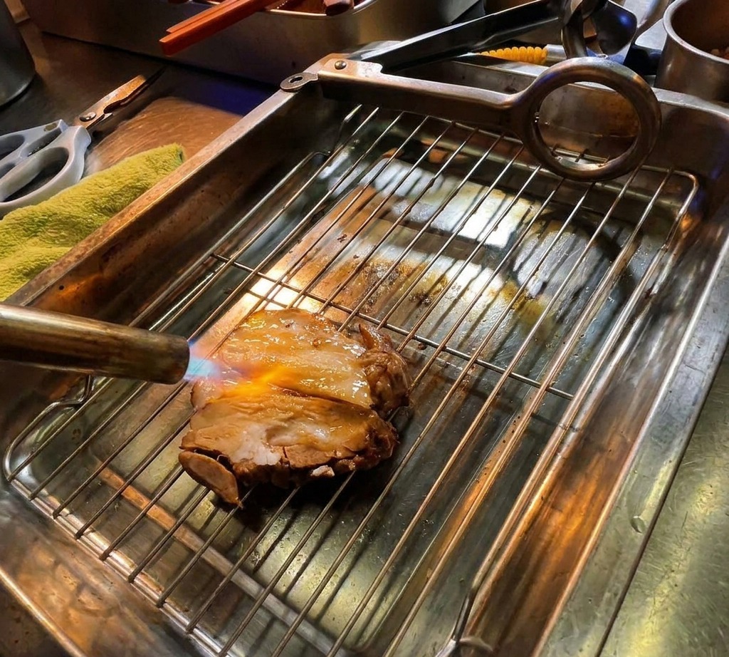 Un soplete de cocina sellando una rodaja gruesa de chashu de cerdo marinado sobre una rejilla metálica, con llamas visibles dorando la carne.