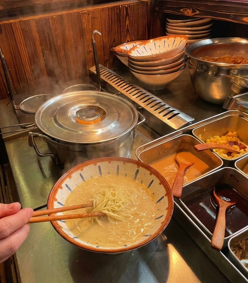 Una persona usando palillos de madera para levantar fideos ramen cocidos y plegarlos en un tazón de caldo cremoso.